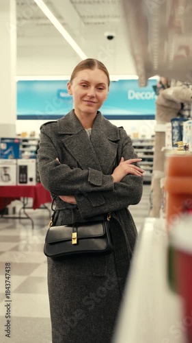 A young woman looks at the camera, wearing a gray coat and carrying a black handbag, with her arms crossed in a clothing store.