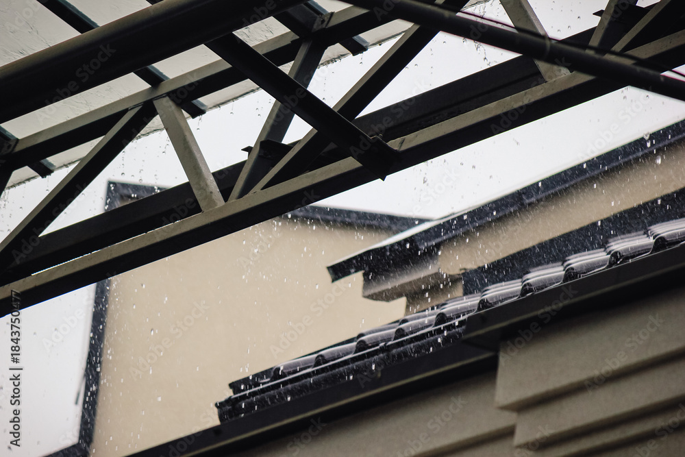 Obraz premium Heavy rain pouring off a black tiled roof and metal gutter below a modern, transparent canopy structure