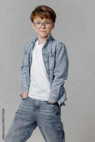 Handsome young man in a denim jacket and glasses poses with his hands in his pockets, radiating confidence and style against an isolated neutral background.