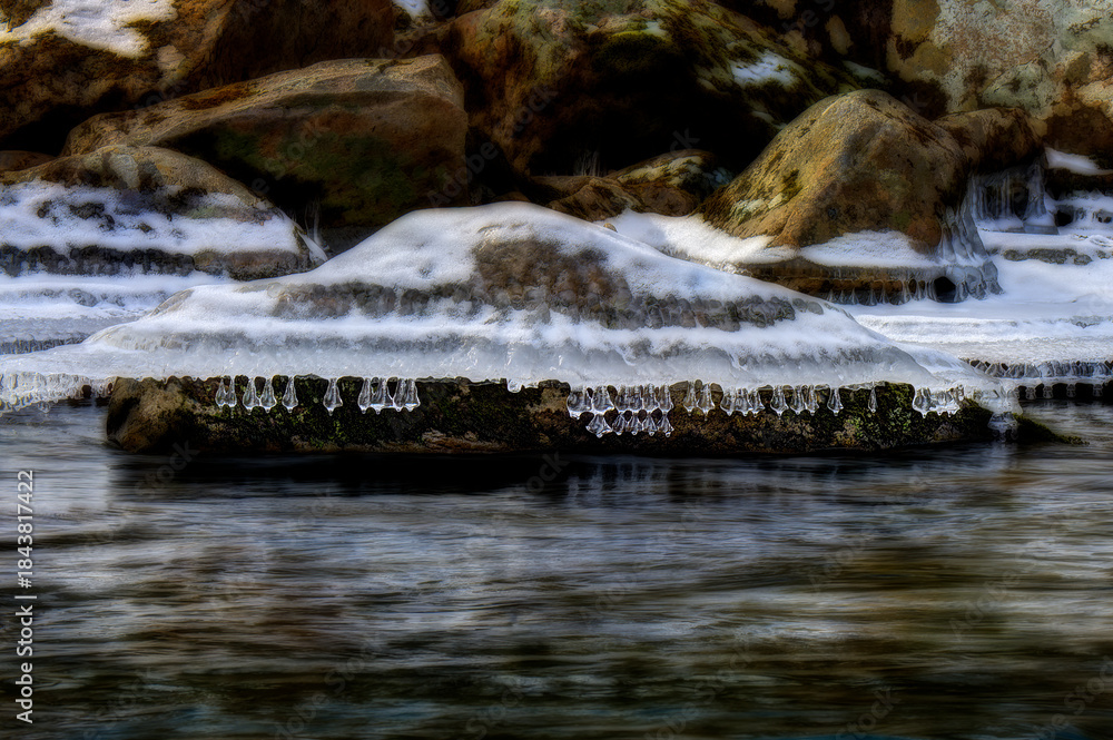 Obraz premium Icicles hang over a flowing river, capturing the contrast between the stillness of the ice and the movement of the water. The scene evokes a sense of tranquility and cold beauty.