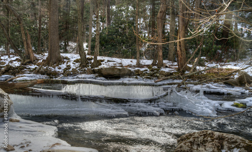 Icicles hang over a flowing stream in a snowy forest. Sunlight filters through the trees, creating a serene, winter atmosphere.