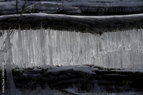 Icicles are forming along the edge of a log, creating a natural curtain of ice. The contrast between the dark wood and bright ice is striking.