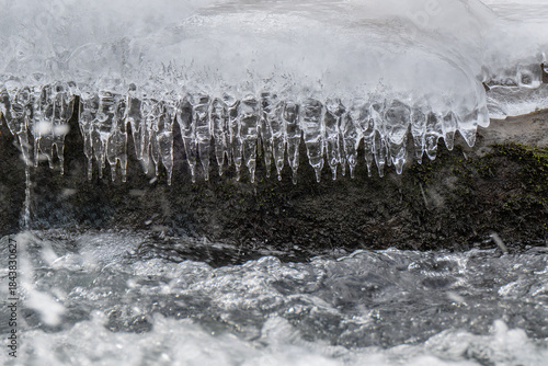 Icicles hang delicately from a snow-covered rock as a stream flows rapidly below. The contrast between the icy formations and the moving water creates a dynamic scene.