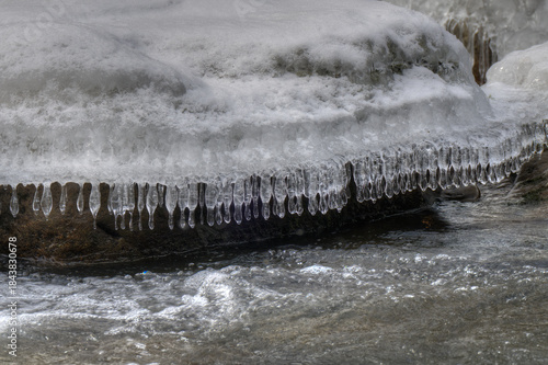 Icicles hang delicately from a snow-covered rock as a stream flows rapidly below. The contrast between the icy formations and the moving water creates a dynamic scene.