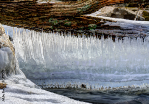 A serene winter scene featuring a fallen tree trunk suspended over a partially frozen stream. Long, delicate icicles hang from the underside of the trunk, forming a shimmering curtain 