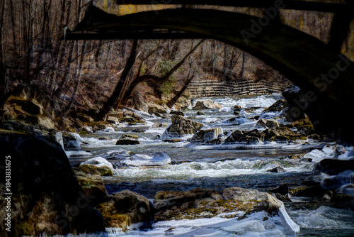 A scenic winter view of a rocky stream rushing through a forested landscape. Whitewater rapids cascade over ice-covered rocks, while leafless trees line the banks in quiet stillness. 