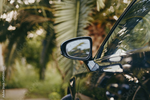 Car side mirror of a black car parked by palm trees in a tropical outdoor travel scene, glossy reflection and summer vibe closeup suggesting a leisure road trip vacation.