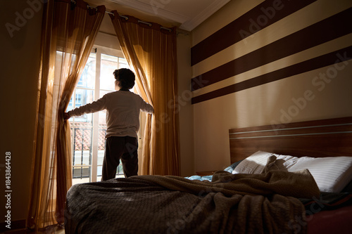 Person Opening Curtains in Sunlit Bedroom by Balcony Door With Striped Wall