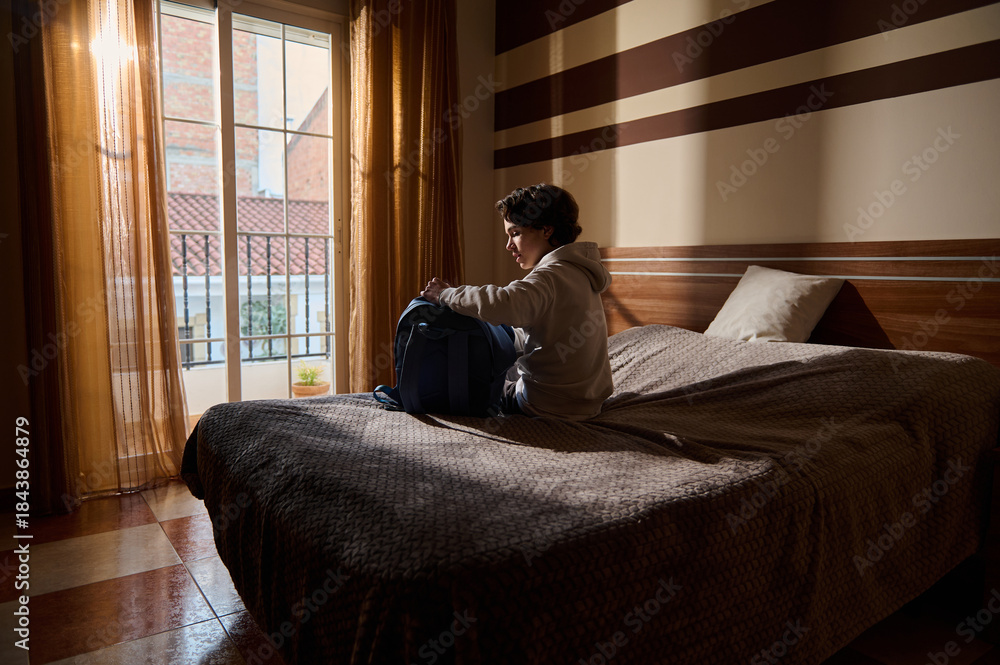 Fototapeta premium Young Man Packing Backpack on Bed in a Sunlit Hotel Room by a Balcony Window