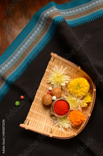 A festive Makar Sankranti set up featuring a bamboo basket holding marigold, til-gul laddus, halwa and kumkum, placed on a black traditional saree.