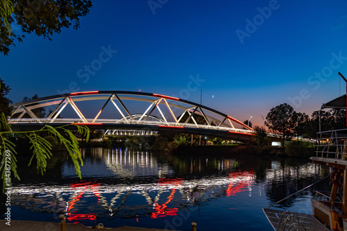 Fototapeta Naklejka Na Ścianę i Meble -  Colorfully illuminated arched bridge over the Manavgat River at night, perfectly reflected in the water with a crescent moon above. A scenic urban nightscape. Manavgat, Turkey, Mediterranean.