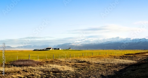 Farmhouse in Iceland. Single white farmhouse in the distance, grassy field in foreground and snow topped mountains and glaciers in the background, on a bright day, with a blue sky. Lonely, quiet mood.