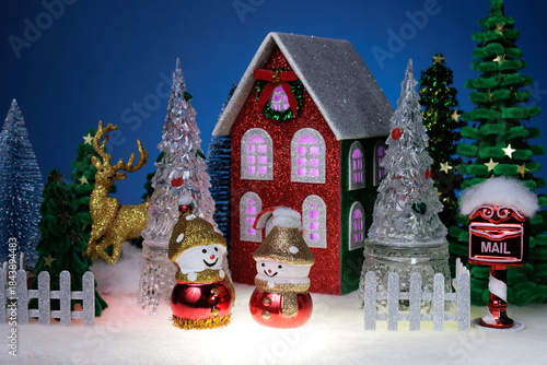 Two identical snowmen among glass Christmas trees near a red Christmas house surrounded by a fence