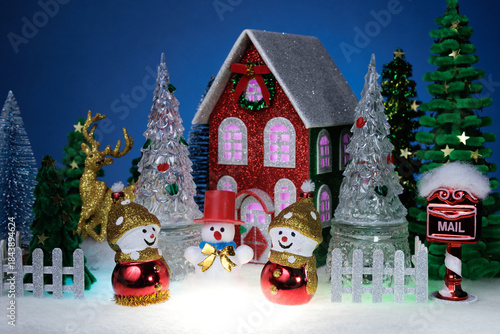 Three snowmen among glass Christmas trees near a red Christmas house surrounded by a fence