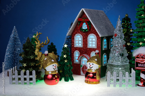 Two identical snowmen among the fir trees near a red Christmas house surrounded by a fence