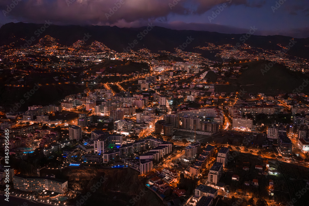 Fototapeta premium Aerial twilight view of Funchal, Madeira shows mid rise buildings, terraced hills, winding lit roads, dark mountains, low clouds, and evening activity with blue lit accents.