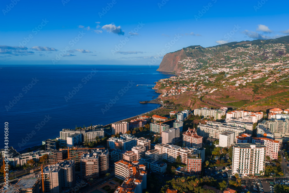 Fototapeta premium Aerial view of Funchal, Madeira, Portugal, with terraced hills, high rise hotels by the promenade, a curving bay, and late day light over deep blue Atlantic waters.