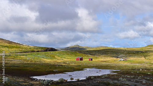Traditional red cabins in remote mountain landscape featuring rolling green hills, dramatic clouds and a small pond. Vestland, Norway, travel Europe
