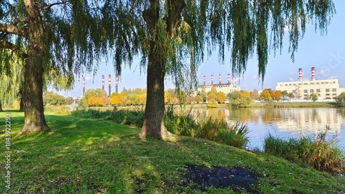 Willows and other trees with yellow leaves grow on the banks of the river, which are reflected in the water. On the opposite shore there are administrative buildings and factory pipes. Sunny autumn