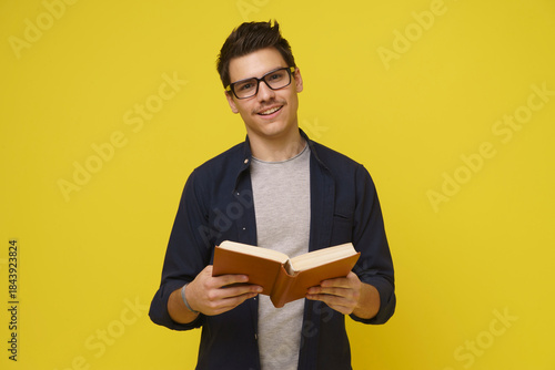 A young man, wearing glasses and a blue shirt, stands against a bright yellow background, holding a book open and looking directly at the camera with a smile