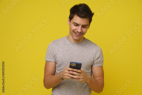 A young Asian man, dressed in a casual grey shirt, smiles brightly as he scrolls through his smartphone, seemingly captivated by the content on his screen