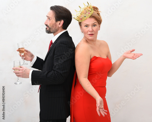 A man in a suit and a woman in a red dress, wearing a crown, stand back-to-back, each holding a glass of champagne. The woman gestures with her left hand, while the man appears to be looking away