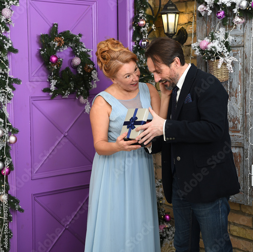 A woman in a blue dress smiles as she presents a gift to a man in a suit. The scene is set against a purple door decorated with a wreath and festive greenery