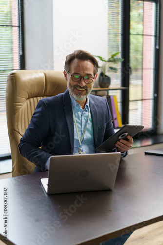 A professional man sits at a modern desk, using a laptop and tablet in a well-lit office. Sunlight streams through large windows, illuminating his focused expression