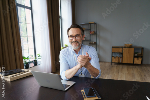 A middle-aged man sits at a sleek desk in a well-lit home office, smiling and clapping his hands after a successful virtual meeting. Plants adorn the room, adding tranquility