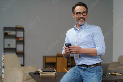 A cheerful middle-aged man interacts with his smartphone, seated by a stylish wooden table in a contemporary office space. Bright light enhances the welcoming atmosphere