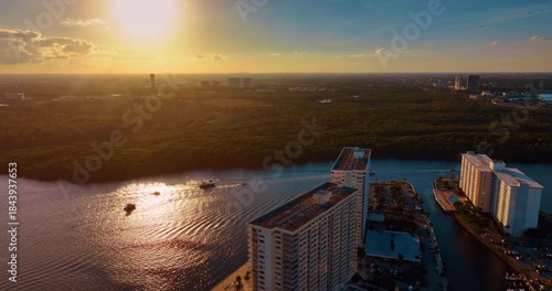 Vessels move by the waterscape dazzled by the setting sun. Lush greenery covers the panorama of the other bank. Aerial view.