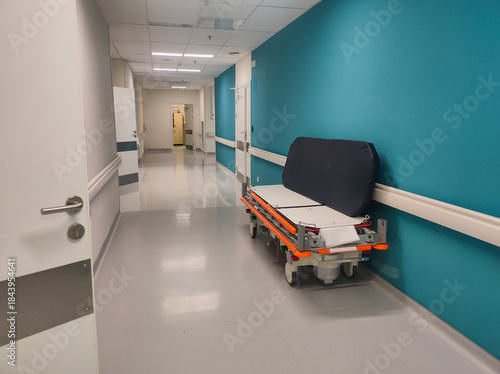 Empty gurney standing in a long, modern hospital corridor with bright blue accent walls and closed doors, representing medical care, emergencies, and the process of healing.