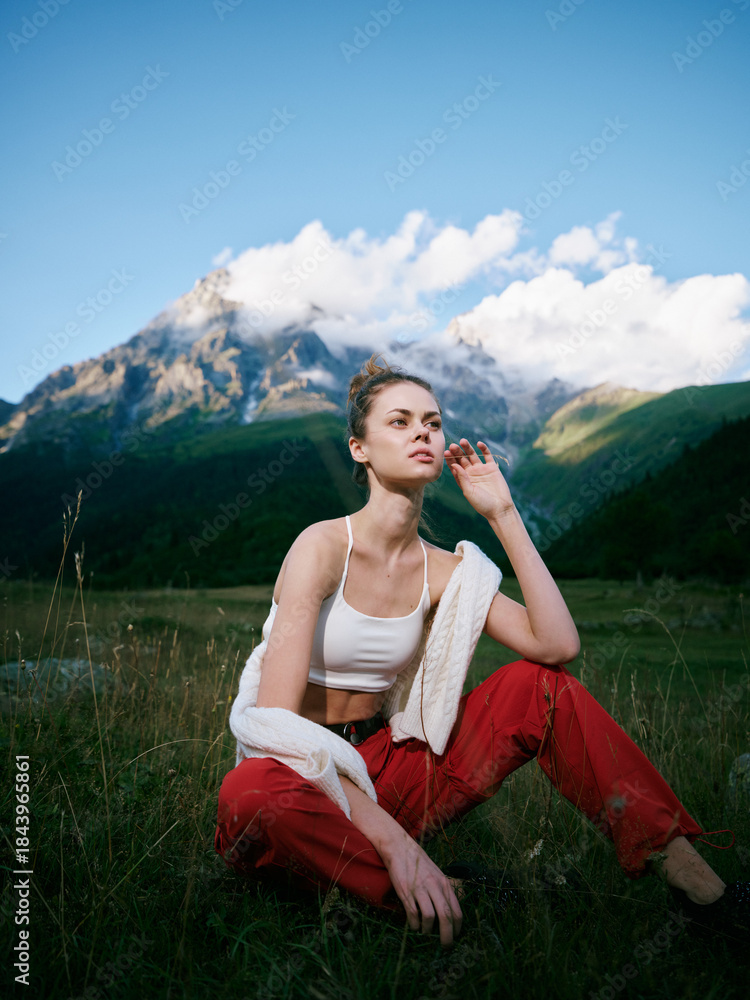 Fototapeta premium Woman sitting on a grassy field with a towel draped over her shoulder, casual white top and red pants, framed by a mountain landscape under a bright blue sky.