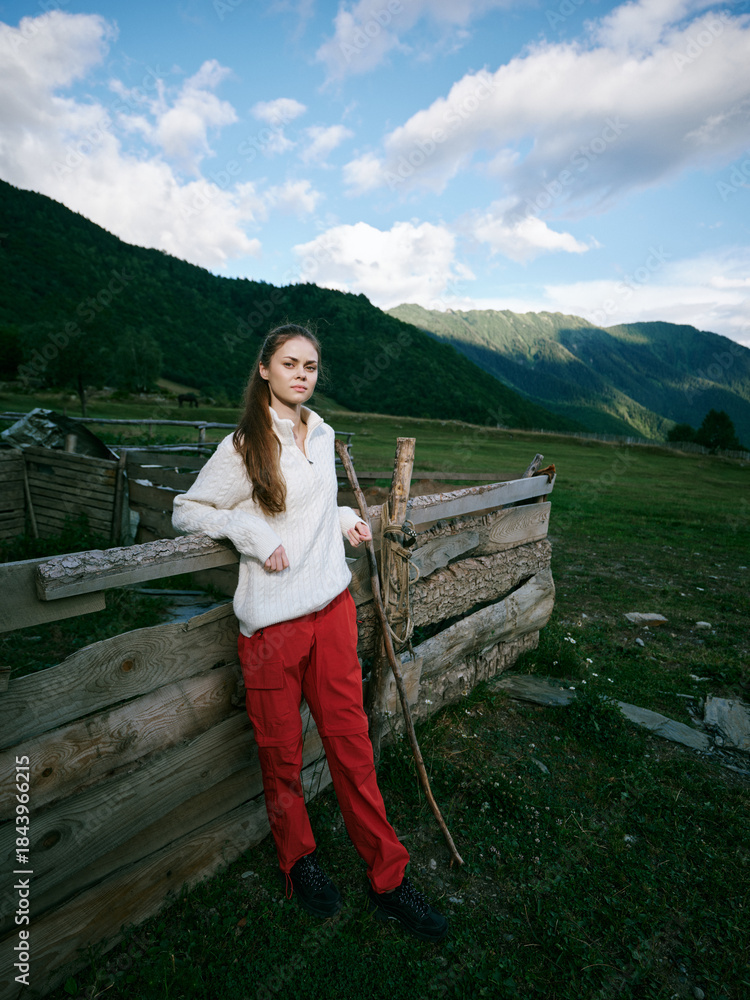 Fototapeta premium Young girl leaning on a rustic wooden fence in a mountainous countryside, wearing a white sweater and red pants, exploring open pasture beneath a bright blue sky.