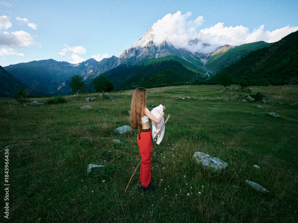Fototapeta premium Mountains, meadow, hiker, outdoors, expansive landscape with rolling field and distant peaks, a figure in red pants holding a jacket, capturing a peaceful moment in nature
