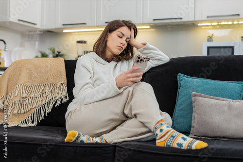 Young woman at home scrolls news on phone feels boredom fatigue stress low energy and low motivation. Caucasian girl on sofa swipes feed frowns sighs feels burnout mood and puts phone down for pause