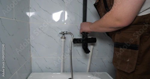 A professional plumber in uniform installs a modern black faucet and shower system. The plumber connects a flexible hose to a water pipe near a tiled wall during a home renovation.