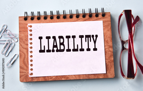 LIABILITY - word on a white sheet of paper on the background of a notebook, glasses and paper clips