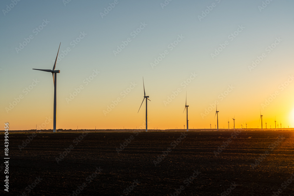 Obraz premium Wind turbines stand across open farmland at sunset, silhouetted against a warm golden sky as the sun drops below the horizon.