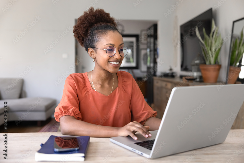 Fototapeta premium Happy beautiful young African businesswoman in elegant glasses working on online freelance project from home, typing on laptop at workplace table, smiling, using wireless technology
