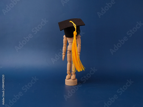 The image features a wooden artist's mannequin wearing a miniature graduation cap and yellow tassel on a blue background