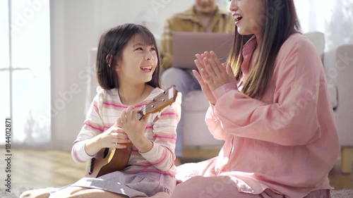 Mother teaching her daughter to play ukulele at home while father works.