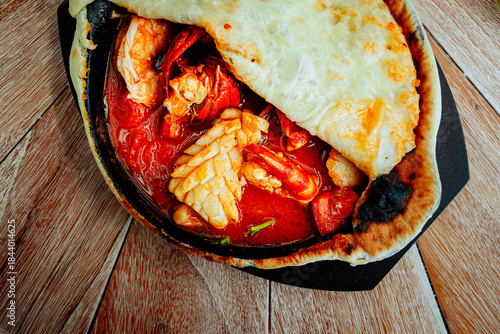 Fotografija A seafood stew en croute, featuring prawns, squid, and crab, simmers in a dark cooking vessel atop a rustic wooden surface