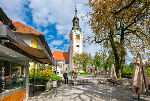 Fotografija Cafes and shops in a small park space under the Church of the Assumption of Mary on Bled Island, on the lake at Bled Slovenia