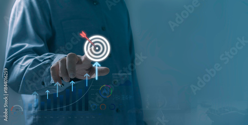 Business growth and target success concept shown by a professional pointing at a digital bullseye icon with rising arrows and analytics charts in a modern data-driven environment.