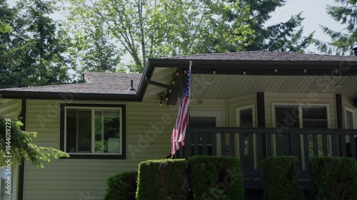 American flag hanging on the porch of a residential house surrounded by green trees. Quiet suburban neighborhood scene showing everyday life, home exterior, and national symbol.