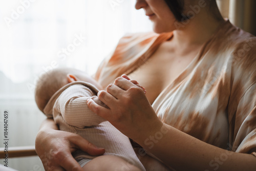 A mother sits in a wooden chair, gently breastfeeding her baby in a warm and softly lit room. Sunlight filters through the curtains, creating a peaceful atmosphere.