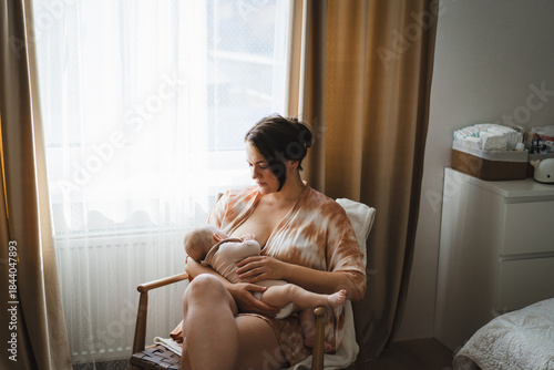 A mother sits in a wooden chair, gently breastfeeding her baby in a warm and softly lit room. Sunlight filters through the curtains, creating a peaceful atmosphere.