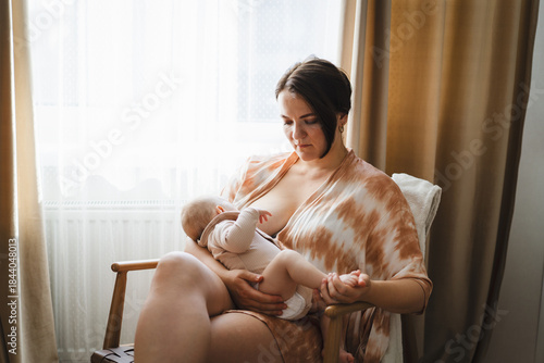 A mother sits in a wooden chair, gently breastfeeding her baby in a warm and softly lit room. Sunlight filters through the curtains, creating a peaceful atmosphere.