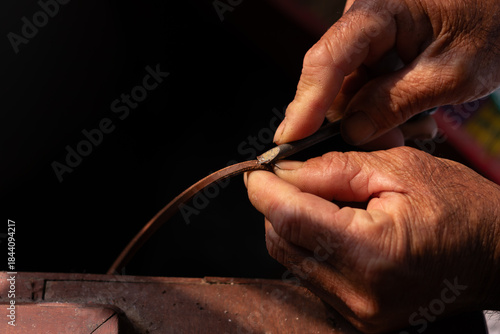 Close up of artisan's hand using a cutting tool to trim a brown leather strip. Handmade craftsmanship and precise leather working process.